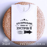 Футболка був нормальний поки не зустрів її, біла футболка, складена на плетеній кошику, фон світло-сірий, принт чорного кольору з текстом та стрілкою, видна етикетка на комірці.