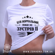 Футболка був нормальний поки не зустрів її, біла футболка, складена на поверхні, фон світло-сірий, принт чорного кольору з текстом та стрілкою, на комірці видна етикетка.
