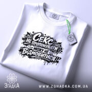 Футболка секс не пропонувати, біла, складена на поверхні, ракурс зверху, фон світло-сірий, принт чорний з текстом і графічними елементами, з етикеткою.