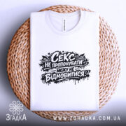 Футболка секс не пропонувати, біла, складена на плетеній основі, ракурс зверху, фон світло-сірий, принт чорний з текстом і графічними елементами.