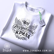Футболка час розправляти крила, біла, складена на поверхні, видно деталі принта з крилами та написом, на фоні світлого кольору, з зеленим ярликом, що підкреслює стиль.