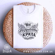 Футболка час розправляти крила, біла, складена на плетеній кошику, принт з крилами та написом, фон світлий, що підкреслює текстуру тканини та стиль оформлення.