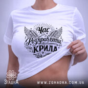 Футболка час розправляти крила, біла, складена на поверхні, видно деталі принта з крилами та написом, на фоні світлого кольору, з зеленим ярликом, що підкреслює стиль.