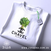 Футболка Chavel Bazar білого кольору, складена на поверхні, ракурс зверху, фон світло-фіолетовий, принт з зображенням пучка шпинату та логотипом CHAVEL BAZAR під ним, з біркою на шнурку.