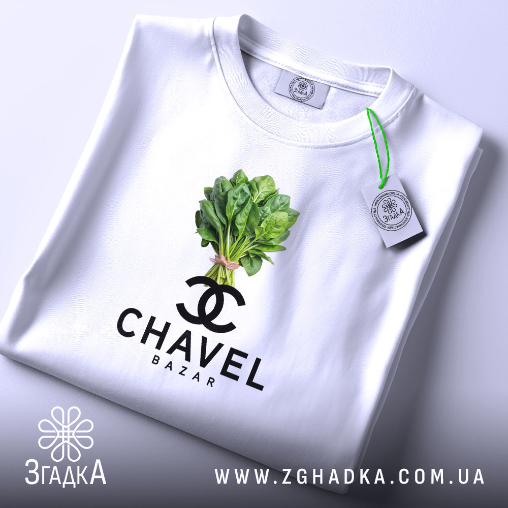 Футболка Chavel Bazar білого кольору, складена на поверхні, ракурс зверху, фон світло-фіолетовий, принт з зображенням пучка шпинату та логотипом CHAVEL BAZAR під ним, з біркою на шнурку.