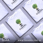 Футболка Chavel Bazar білого кольору, на моделі з коротким рукавом, ракурс спереду, фон світло-фіолетовий, принт з зображенням пучка шпинату та логотипом CHAVEL BAZAR під ним.