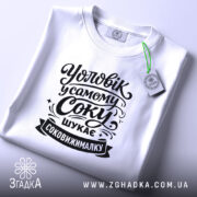 Футболка чоловік у самому соку, біла, з верхнього ракурсу, на фоні світло-фіолетового кольору, з чорним принтом, що містить текст "Уловік у самому соку шукає соковижималку", з етикеткою зеленого кольору.