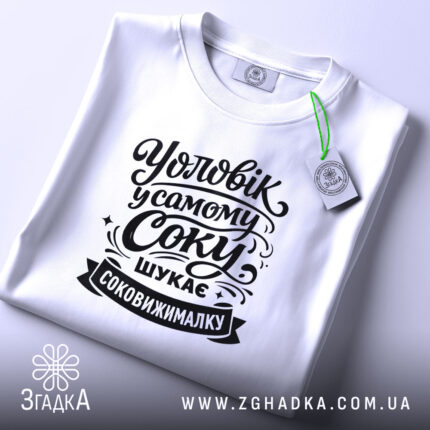 Футболка чоловік у самому соку, біла, з верхнього ракурсу, на фоні світло-фіолетового кольору, з чорним принтом, що містить текст "Уловік у самому соку шукає соковижималку", з етикеткою зеленого кольору.