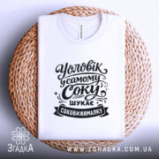 Футболка чоловік у самому соку, біла, з верхнього ракурсу, на фоні світло-сірого кольору, з чорним принтом, що містить текст "Уловік у самому соку шукає соковижималку", розташована на плетеній основі.