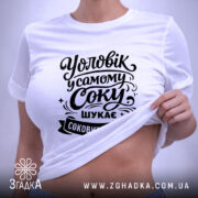 Футболка чоловік у самому соку, біла, з верхнього ракурсу, на фоні світло-фіолетового кольору, з чорним принтом, що містить текст "Уловік у самому соку шукає соковижималку", з етикеткою зеленого кольору.