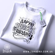 Футболка дарую щастя щодня, біла, на плоскій поверхні, фон світло-сірий, принт чорного кольору з написом та елементами декору, з етикеткою на шнурку.