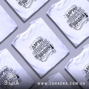 Футболка дарую щастя щодня, біла, на моделі з легким ракурсом, фон ніжно-фіолетовий, принт чорного кольору з написом та елементами декору.