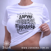 Футболка дарую щастя щодня, біла, на плоскій поверхні, фон світло-сірий, принт чорного кольору з написом та елементами декору, з етикеткою на шнурку.