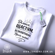 Футболка ділюся щастям. Біла футболка, складена на столі, з принтом чорного кольору, на фоні світло-сірого кольору. Принт містить текст з позитивним посланням і малюнком сонця, з етикеткою на зеленій стрічці.