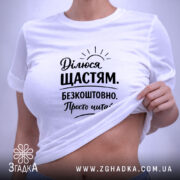 Футболка ділюся щастям. Біла футболка, складена на столі, з принтом чорного кольору, на фоні світло-сірого кольору. Принт містить текст з позитивним посланням і малюнком сонця, з етикеткою на зеленій стрічці.