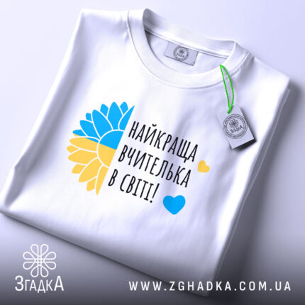 Футболка для найкращої вчительки, білого кольору, складена на плоскій поверхні, фон світло-сірий, принт з жовто-блакитним соняшником та написом 'найкраща вчителька в світі!' у чорному кольорі, на комірці видна етикетка.