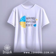 Футболка для найкращої вчительки, білого кольору, підвішена на вішалці, фон світло-сірий, принт з жовто-блакитним соняшником та написом 'найкраща вчителька в світі!' у чорному кольорі, видна форма рукавів.