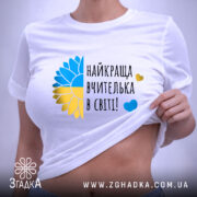 Футболка для найкращої вчительки, білого кольору, складена на плоскій поверхні, фон світло-сірий, принт з жовто-блакитним соняшником та написом 'найкраща вчителька в світі!' у чорному кольорі, на комірці видна етикетка.