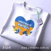 Футболка для вчителя випускників, біла, на плоскій поверхні, з принтом у формі серця, що містить синій і жовтий кольори, з написом 'НАЙКРАЩИЙ ВЧИТЕЛЬ' та квітами, з етикеткою, що звисає з коміра.