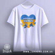 Футболка для вчителя випускників, біла, підвішена на вішалці, з принтом у формі серця, що містить синій і жовтий кольори, з написом 'НАЙКРАЩИЙ ВЧИТЕЛЬ' та квітами, фон світло-сірий.