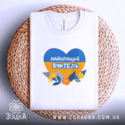 Футболка для вчителя випускників, біла, на плоскій поверхні, з принтом у формі серця, що містить синій і жовтий кольори, з написом 'НАЙКРАЩИЙ ВЧИТЕЛЬ' та квітами, розташована на плетеній основі.