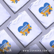 Футболка для вчителя випускників, біла, на моделі, з принтом у формі серця, що містить синій і жовтий кольори, з написом 'НАЙКРАЩИЙ ВЧИТЕЛЬ' та квітами на фоні, модель позує з легким усмішкою на нейтральному фоні.