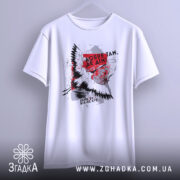 Футболка добре там де дім. Біла футболка підвішена на вішалці, з принтом птаха та текстом. Фон світло-сірий, футболка має простий, але стильний вигляд.