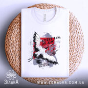 Футболка добре там де дім. Біла футболка, складена на плетеному кошику, з принтом птаха та текстом. Фон світло-сірий, футболка виглядає акуратно та стильно.