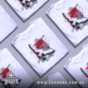 Футболка добре там де дім. Біла футболка на моделі, з принтом на грудях, що містить зображення птаха та текст. Фон світло-фіолетовий, модель позує з легким усмішкою, підкреслюючи стиль футболки.