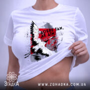 Футболка добре там де дім. Біла футболка, складена на столі, з принтом, що включає птаха та текст. Фон світло-сірий, на футболці видно етикетку з зеленим шнурком.