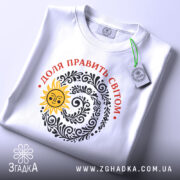 Футболка Доля править світом, біла, на плоскому фоні, з верхнього ракурсу, принт з сонцем та візерунками чорного кольору, текст червоним кольором, етикетка з зеленим шнурком.