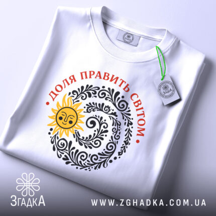 Футболка Доля править світом, біла, на плоскому фоні, з верхнього ракурсу, принт з сонцем та візерунками чорного кольору, текст червоним кольором, етикетка з зеленим шнурком.