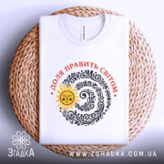 Футболка Доля править світом, біла, на плоскому фоні, з верхнього ракурсу, принт з сонцем та візерунками чорного кольору, текст червоним кольором, розташована на плетеному кошику.