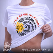 Футболка Доля править світом, біла, на плоскому фоні, з верхнього ракурсу, принт з сонцем та візерунками чорного кольору, текст червоним кольором, етикетка з зеленим шнурком.