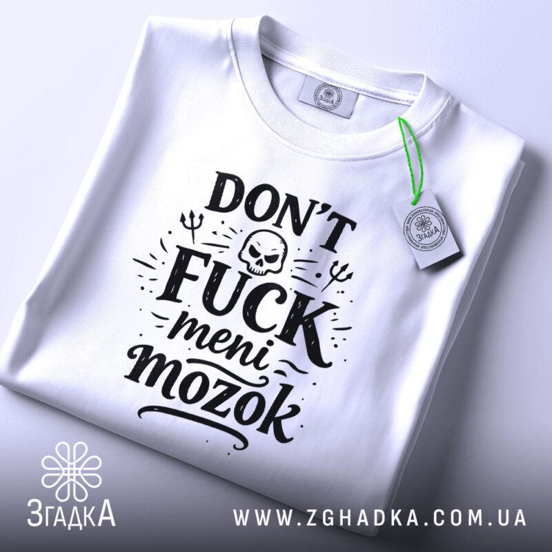 Футболка Don’t Fuck meni mozok, біла, складена на поверхні, ракурс зверху, фон світло-сірий, принт чорного кольору з черепом та декоративними елементами, на ярлику зелена стрічка.