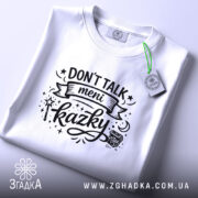Футболка Don’t Talk мені казки білого кольору, складена на поверхні, фон світло-сірий, принт чорного кольору з написом 'Don't Talk meni kazky' та елементами дизайну, на футболці також є етикетка з зеленим шнурком.