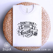 Футболка Don’t Talk мені казки білого кольору, складена на плетеній кошику, фон світло-сірий, принт чорного кольору з написом 'Don't Talk meni kazky' та елементами дизайну, видима етикетка.