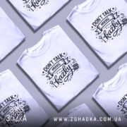 Футболка Don’t Talk мені казки білого кольору, зображена на моделі з коротким рукавом, фон ніжно-фіолетовий, принт чорного кольору з написом 'Don't Talk meni kazky' та декоративними елементами.