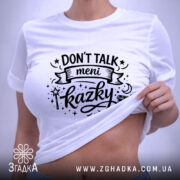 Футболка Don’t Talk мені казки білого кольору, складена на поверхні, фон світло-сірий, принт чорного кольору з написом 'Don't Talk meni kazky' та елементами дизайну, на футболці також є етикетка з зеленим шнурком.
