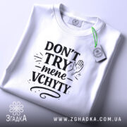Футболка Don’t Try mene vchyty білого кольору, складена на поверхні з нейтральним фоном. Принт чорного кольору з текстом та малюнком руки, розташований по центру, з етикеткою, що звисає з коміра.