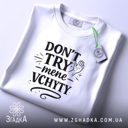 Футболка Don’t Try mene vchyty білого кольору, складена на поверхні з нейтральним фоном. Принт чорного кольору з текстом та малюнком руки, розташований по центру, з етикеткою, що звисає з коміра.