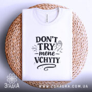 Футболка Don’t Try mene vchyty білого кольору, складена на плетеній кошику, що має натуральний вигляд. Принт чорного кольору з текстом та малюнком руки, розташований по центру, з видимою етикеткою на комірі.