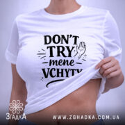 Футболка Don’t Try mene vchyty білого кольору, складена на поверхні з нейтральним фоном. Принт чорного кольору з текстом та малюнком руки, розташований по центру, з етикеткою, що звисає з коміра.