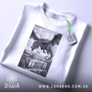 Футболка Дракон над Радою, біла, розкладена на поверхні, злегка під кутом, фон світло-сірий, принт з чорним драконом, що летить над будівлею з українським прапором, на комірці видна етикетка.