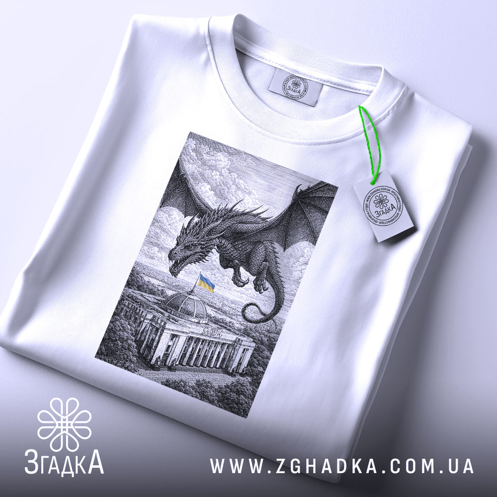 Футболка Дракон над Радою, біла, розкладена на поверхні, злегка під кутом, фон світло-сірий, принт з чорним драконом, що летить над будівлею з українським прапором, на комірці видна етикетка.
