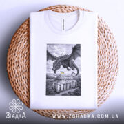 Футболка Дракон над Радою, біла, розкладена на плетеній кошику, фон світло-сірий, принт з чорним драконом, що летить над будівлею з українським прапором, видна етикетка на комірці.