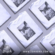 Футболка Дракон над Радою, біла, зображена на моделі, яка позує з легким усмішкою, фон світло-фіолетовий, принт з чорним драконом, що летить над будівлею з українським прапором.