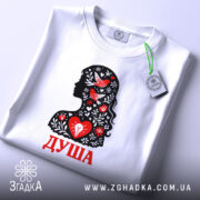 Футболка Душа український стиль, біла, складена на поверхні, фон світло-сірий, принт з силуетом жінки, прикрашеним червоними пташками та квітами, текст 'ДУША' червоним кольором, на етикетці зелена стрічка.