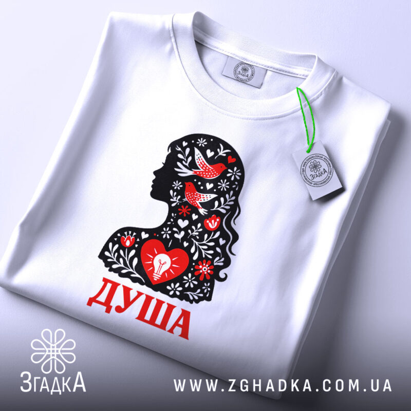 Футболка Душа український стиль, біла, складена на поверхні, фон світло-сірий, принт з силуетом жінки, прикрашеним червоними пташками та квітами, текст 'ДУША' червоним кольором, на етикетці зелена стрічка.