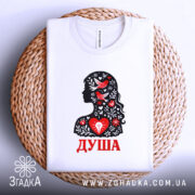 Футболка Душа український стиль, біла, складена на плетеній основі, фон світло-сірий, принт з силуетом жінки, прикрашеним червоними пташками та квітами, текст 'ДУША' червоним кольором.